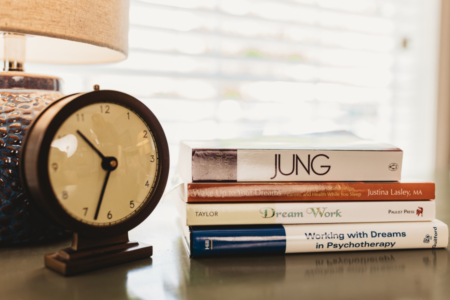 A small clock beside a stack of books on Jung and dream work on a table by a sunlit window.