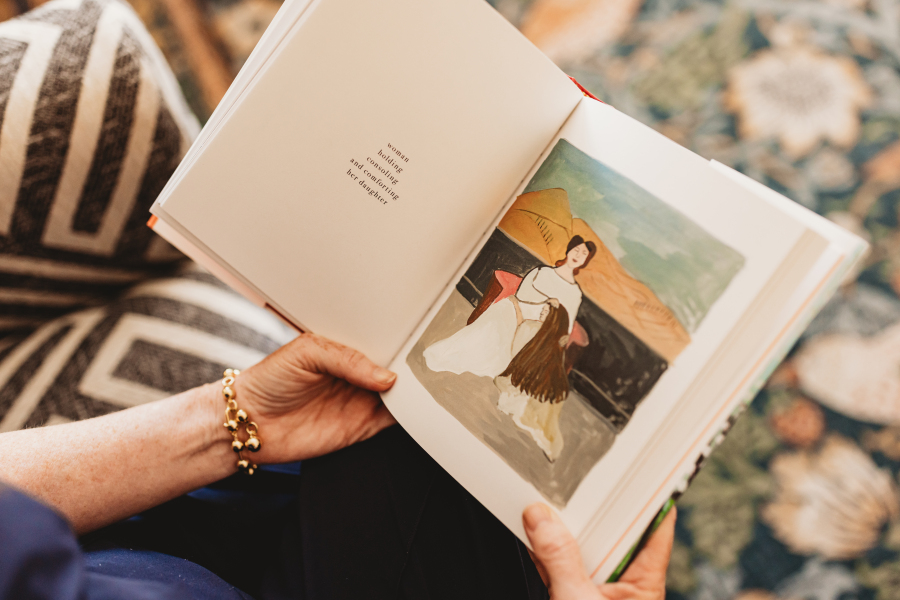 A book open in Lisa Giebelhaus's hands, its page captioned "woman holding, consoling and comforting her daughter" beside a painted illustration.