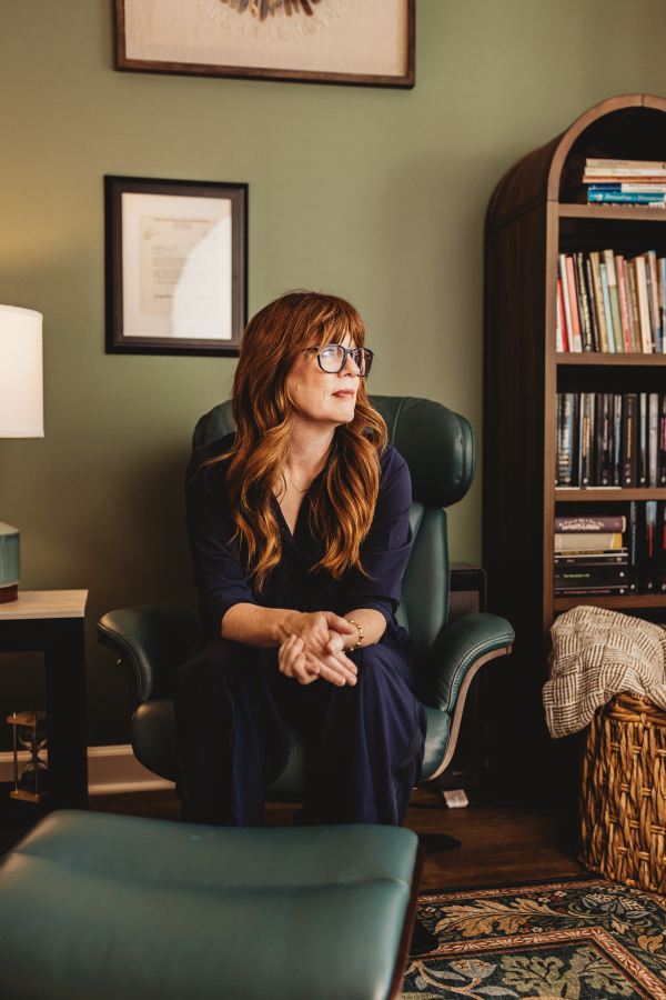 Lisa Giebelhaus sits forward in the green leather chair in her office, hands clasped, looking away toward the window past a framed credential and an arched bookcase